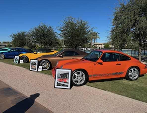 Classic Porsches lined up on a lawn beside framed prints
